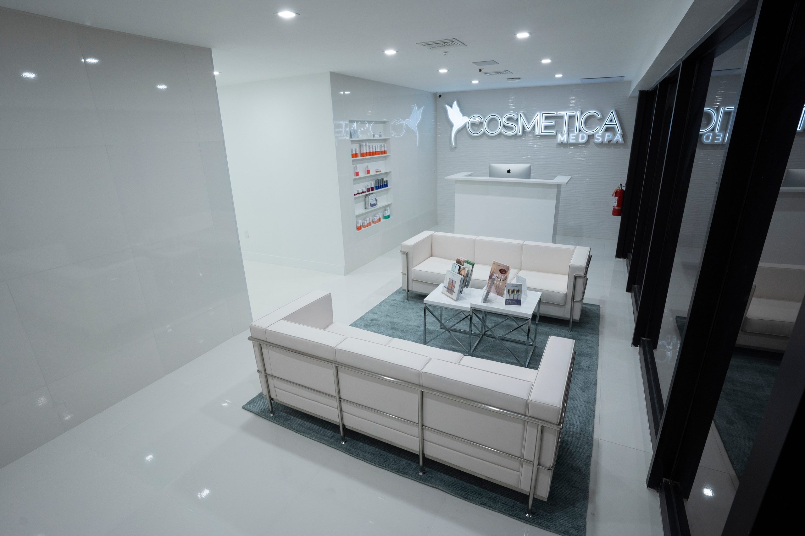 Waiting lounge with white sofas on a teal rug and the reception desk behind at Cosmetica Med Spa