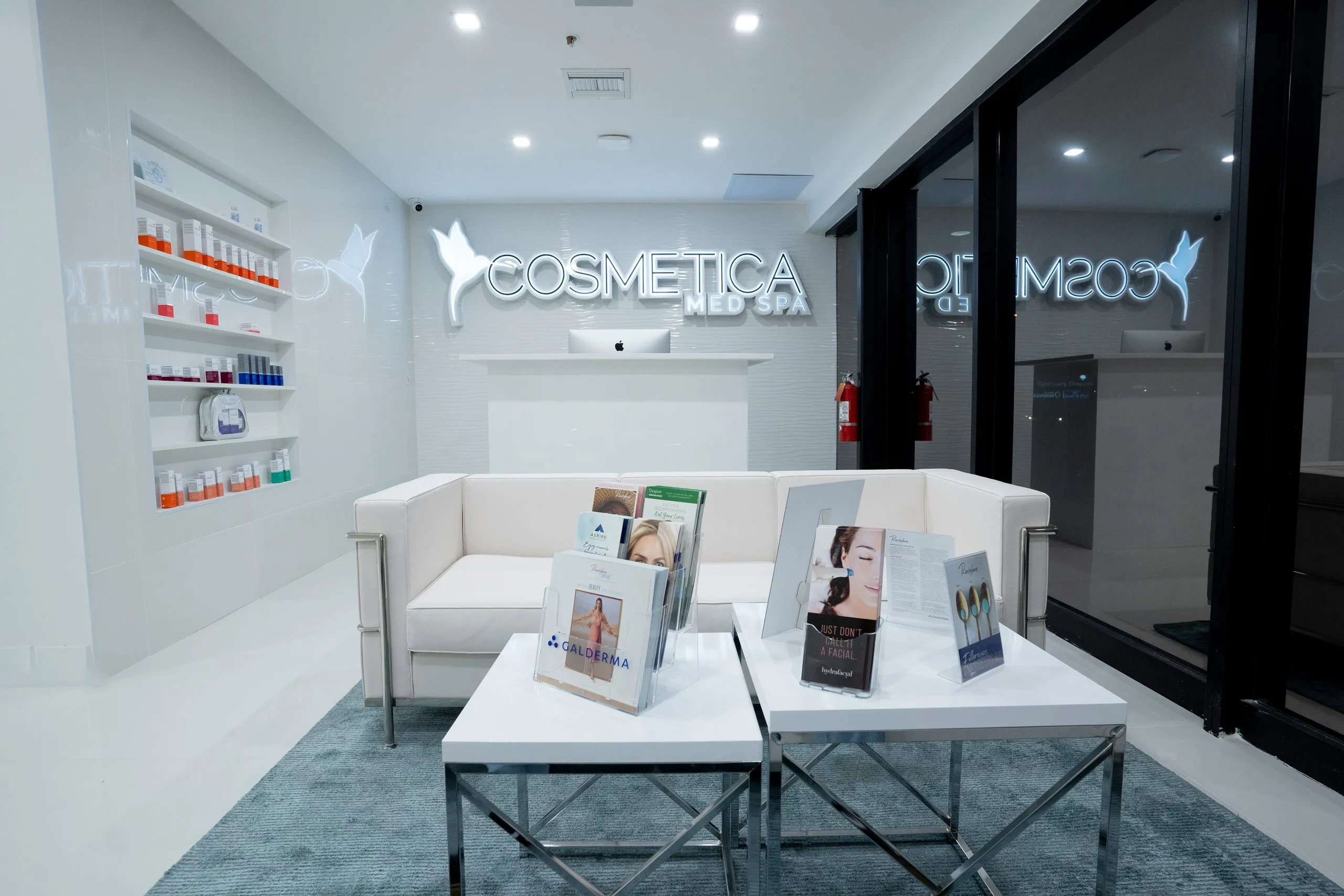 Brochure-topped coffee tables in front of the lit reception logo wall at Cosmetica Med Spa