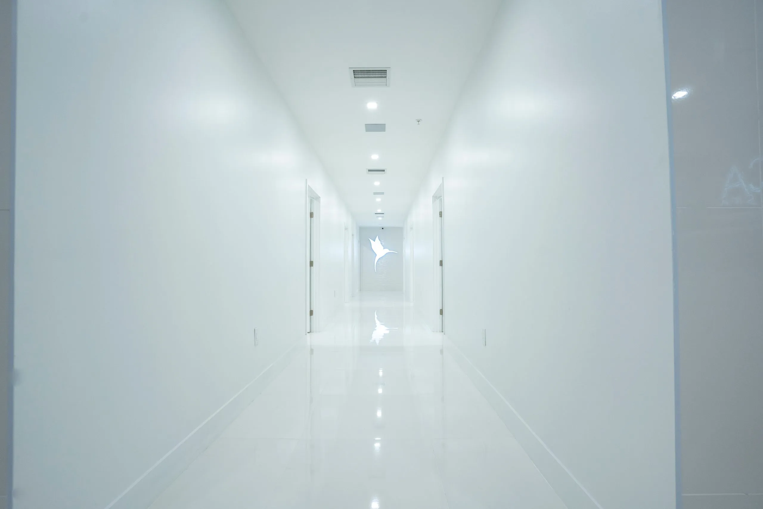 Bright white corridor with recessed lights leading to the hummingbird logo at Cosmetica Med Spa