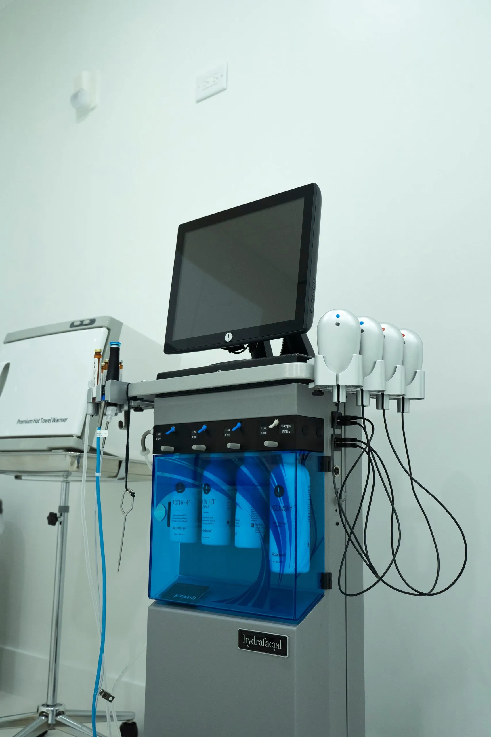 HydraFacial machine with a touchscreen and four handpieces beside a towel warmer at Cosmetica Med Spa