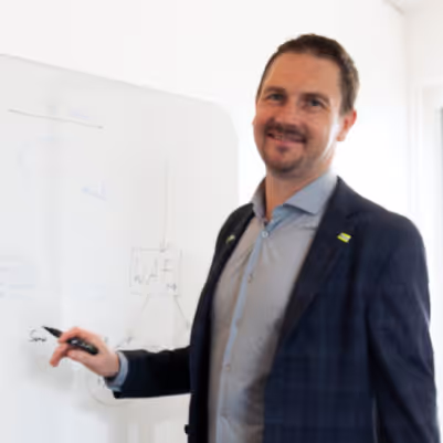 Ein lächelnder Mann in Hemd und Sakko steht vor einem Whiteboard und hält einen Marker.