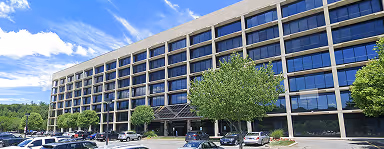 Modern office building with reflective windows, trees, and a parking lot under a blue sky.