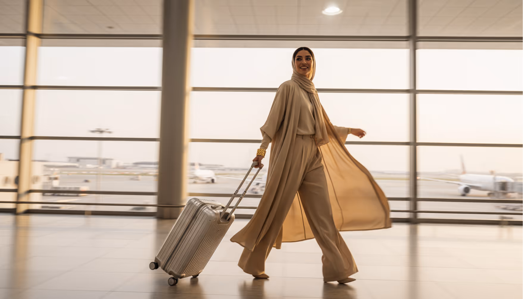 Woman in beige outfit and headscarf walking through airport with rolling suitcase.