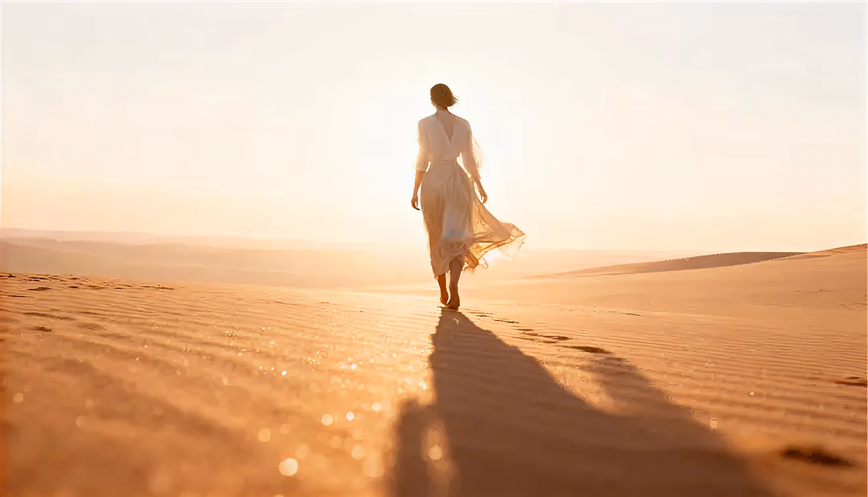 Woman in a long dress walking barefoot on sunlit desert sand with a long shadow.