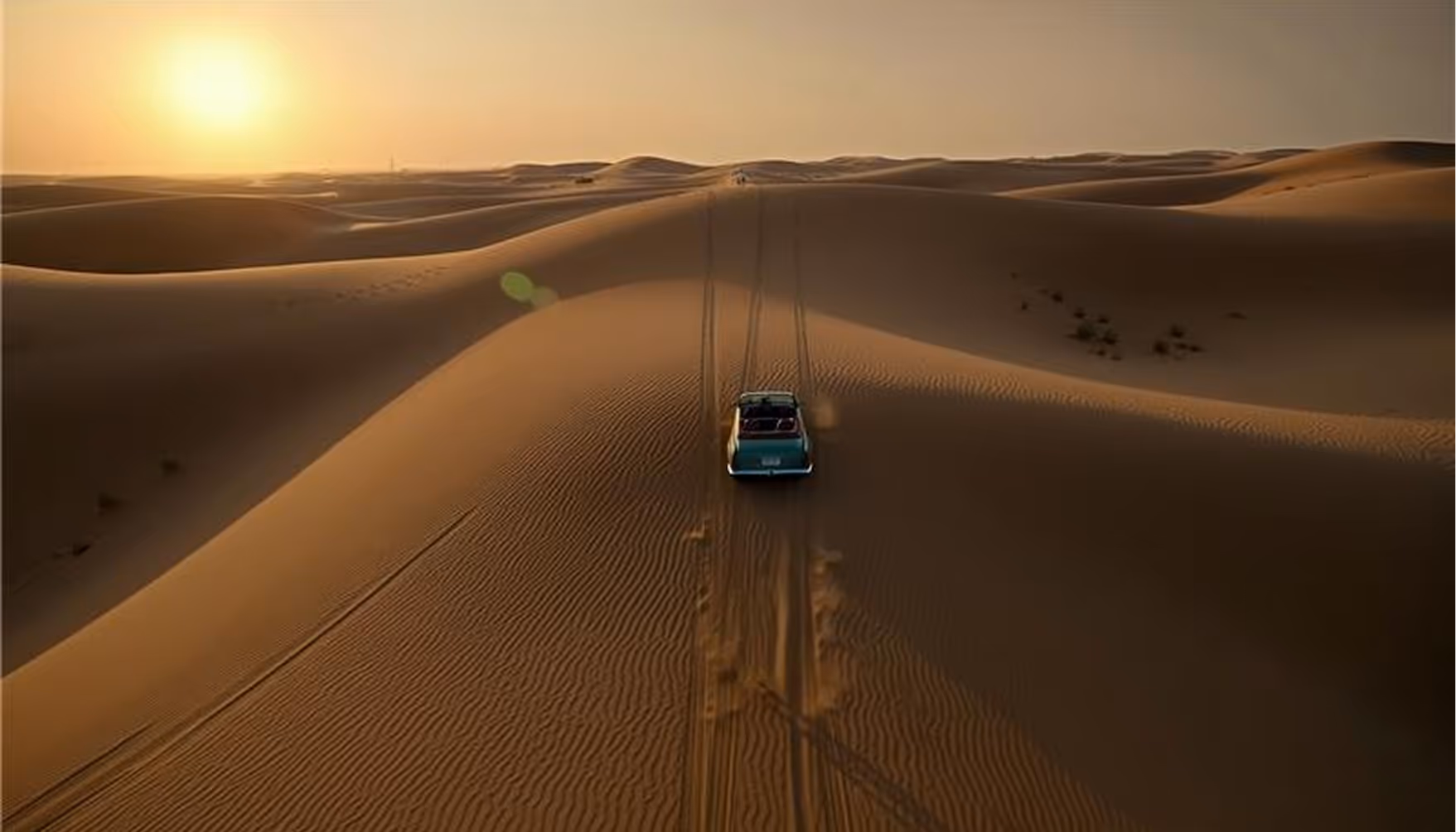 A blue off-road vehicle drives over sand dunes at sunset in a vast desert landscape.