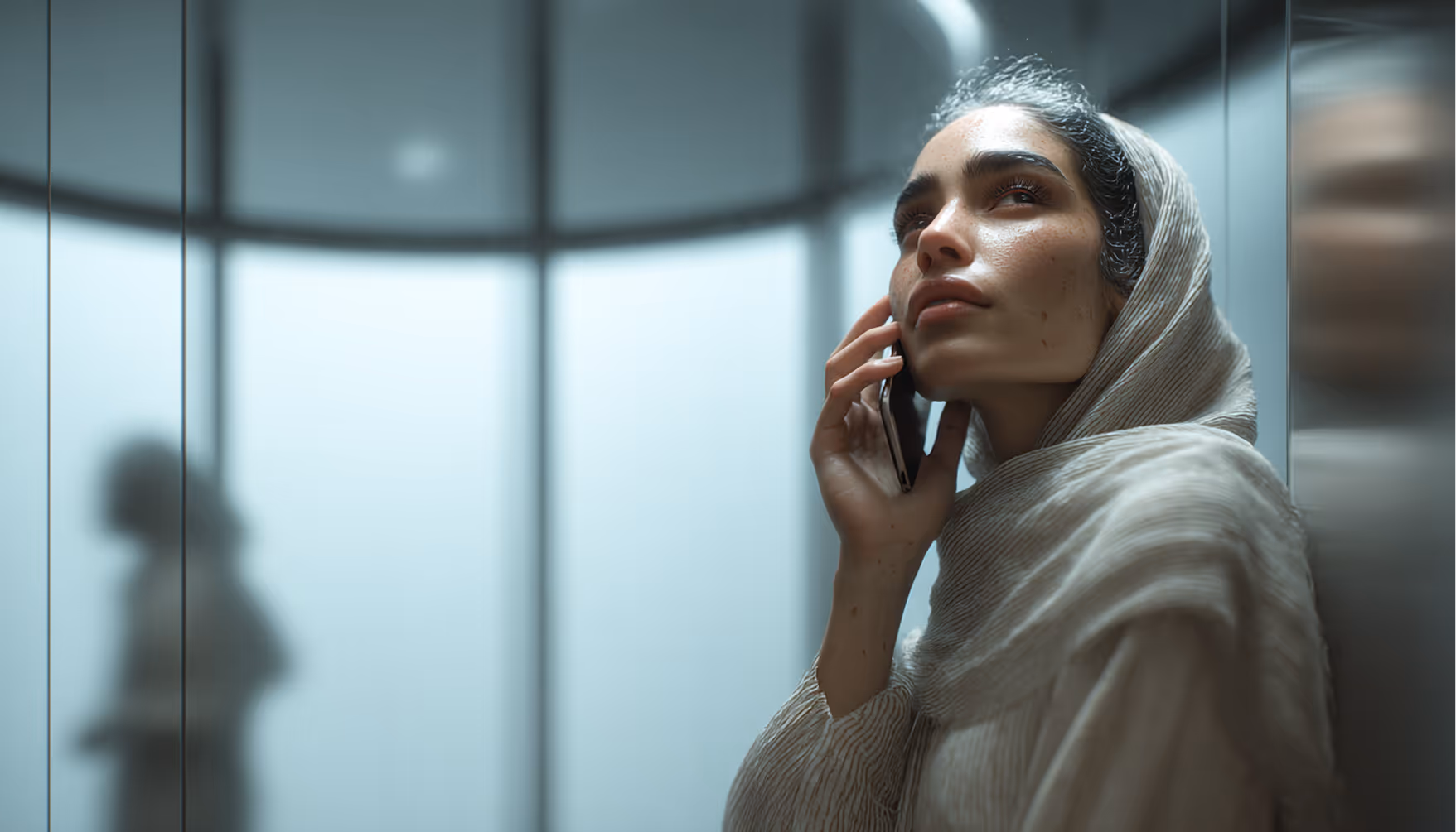 Woman in a light hijab leaning against elevator wall, talking on a phone and looking upward thoughtfully.