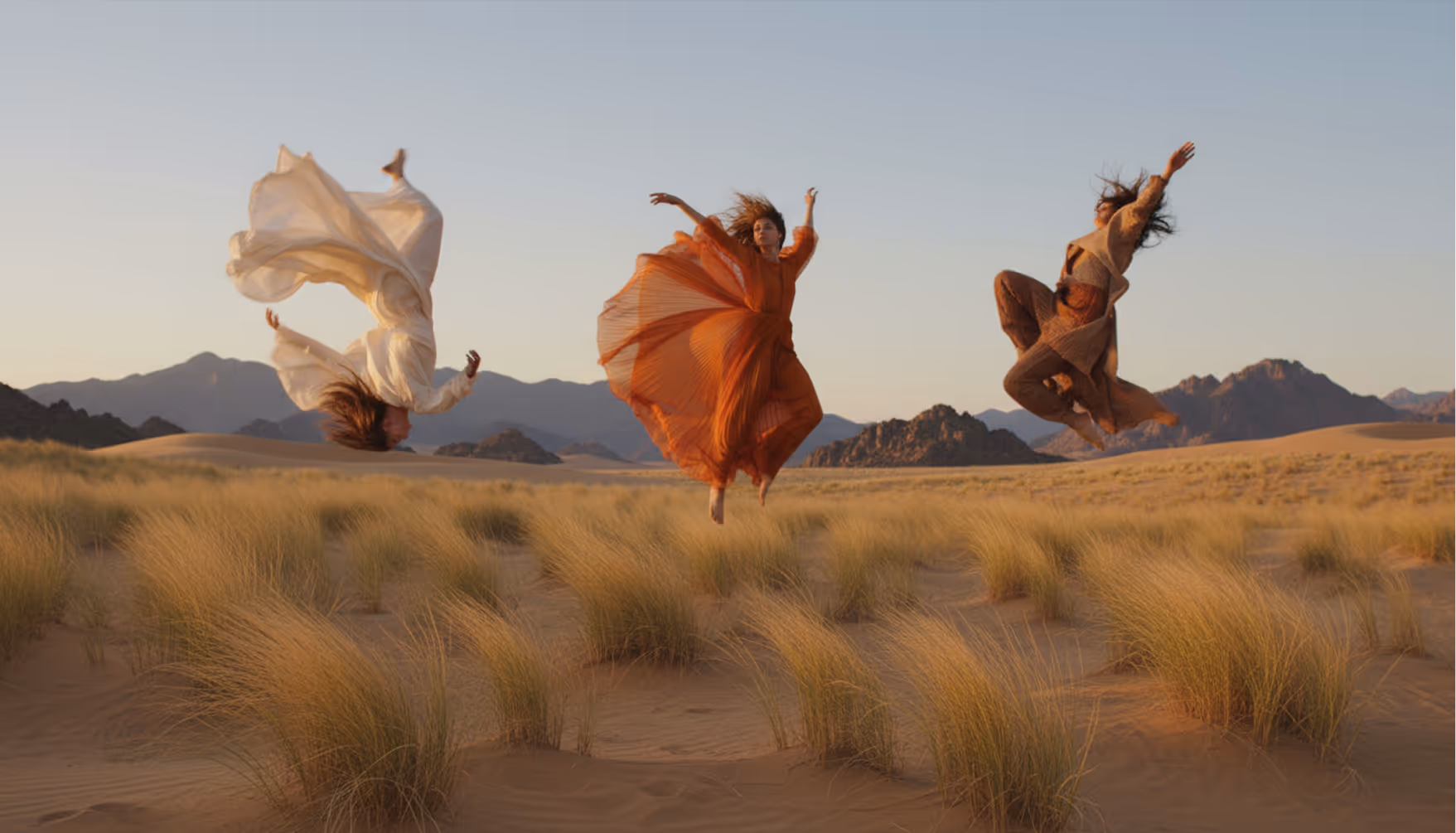 Three women in flowing dresses leap in a desert field with mountains in the background at sunset.