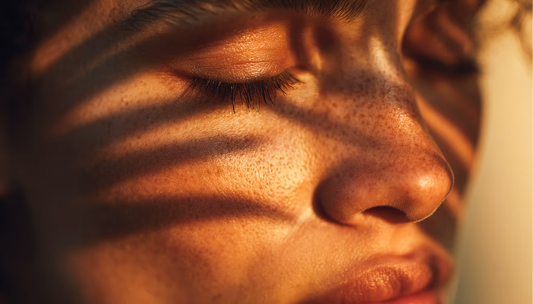 Close-up of a face with closed eyes and shadow stripes across freckled skin in warm light.