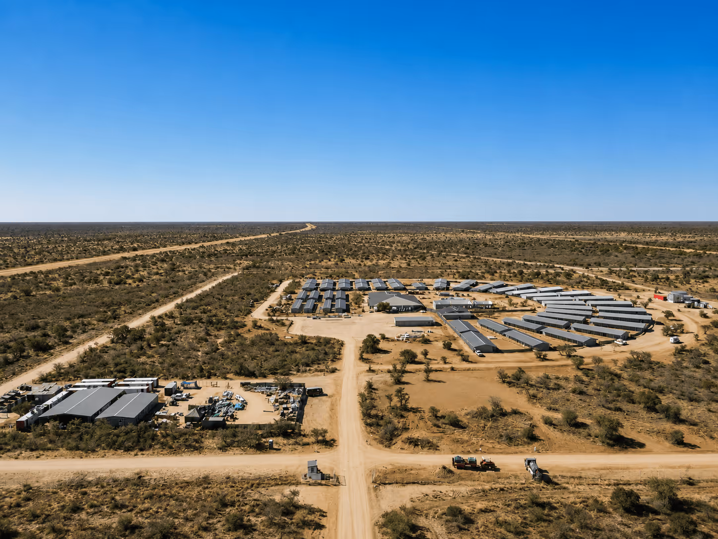 Aerial view of a remote industrial site in a dry, flat landscape with multiple rows of solar panels arranged in a circular formation and nearby storage buildings, surrounded by dirt roads and sparse vegetation under a clear blue sky.