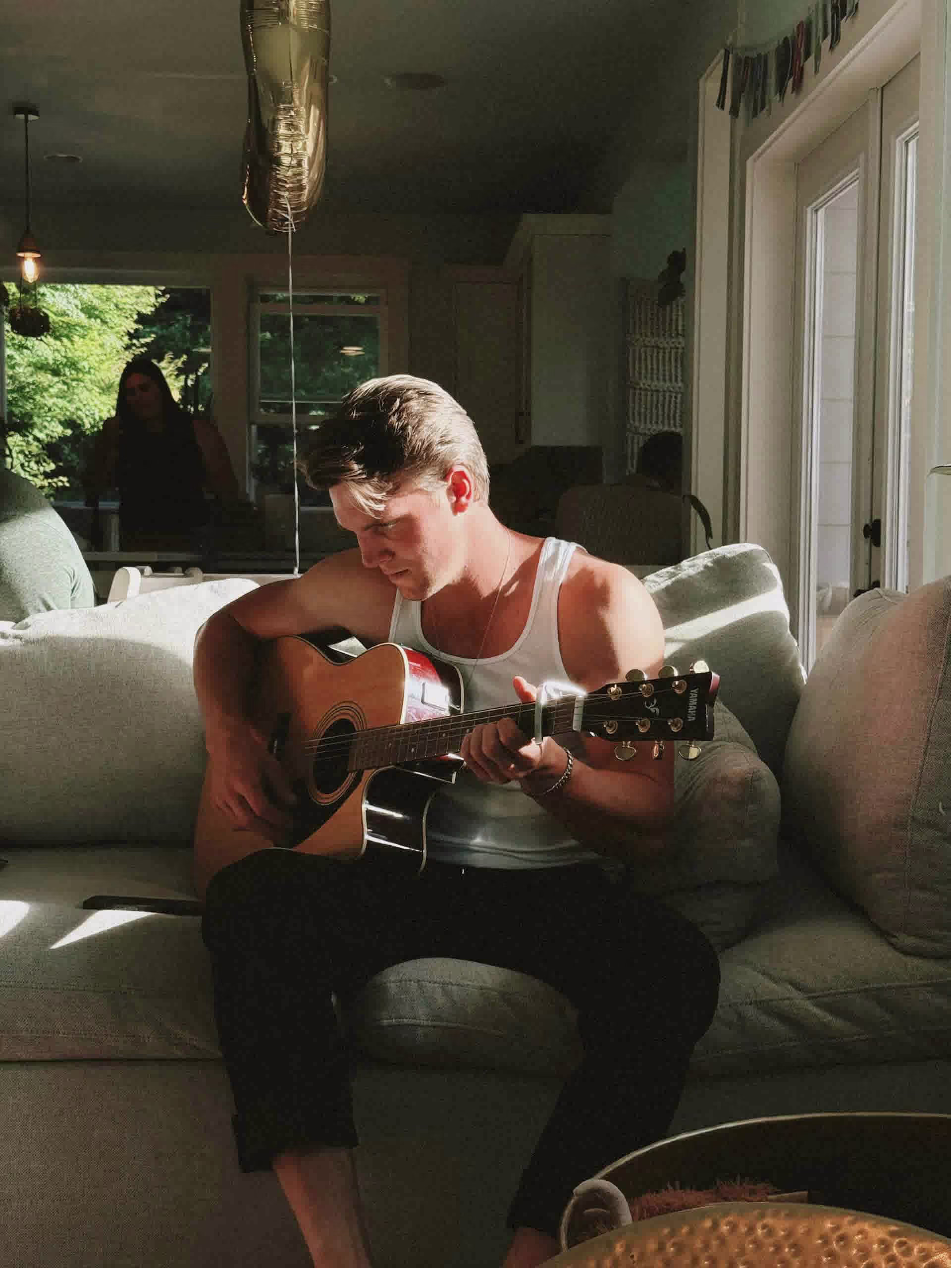 Young man in white tank top sitting on a couch playing an acoustic guitar in sunlight.