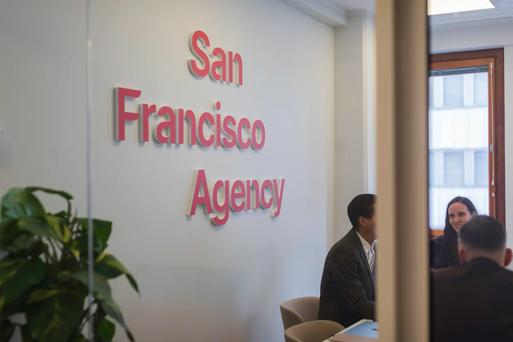 View through a glass wall into the San Francisco Agency office, where three people talk beside the red wall sign