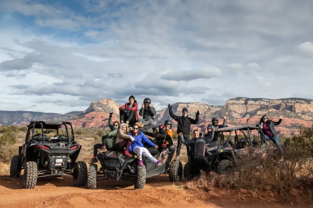 Group of people posing on off-road vehicles on a red dirt track below red rock cliffs