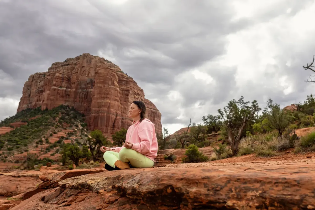 Woman meditating cross-legged on red rock below a tall butte under cloudy skies
