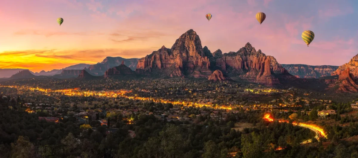 Hot air balloons over red rock peaks and town lights at sunset