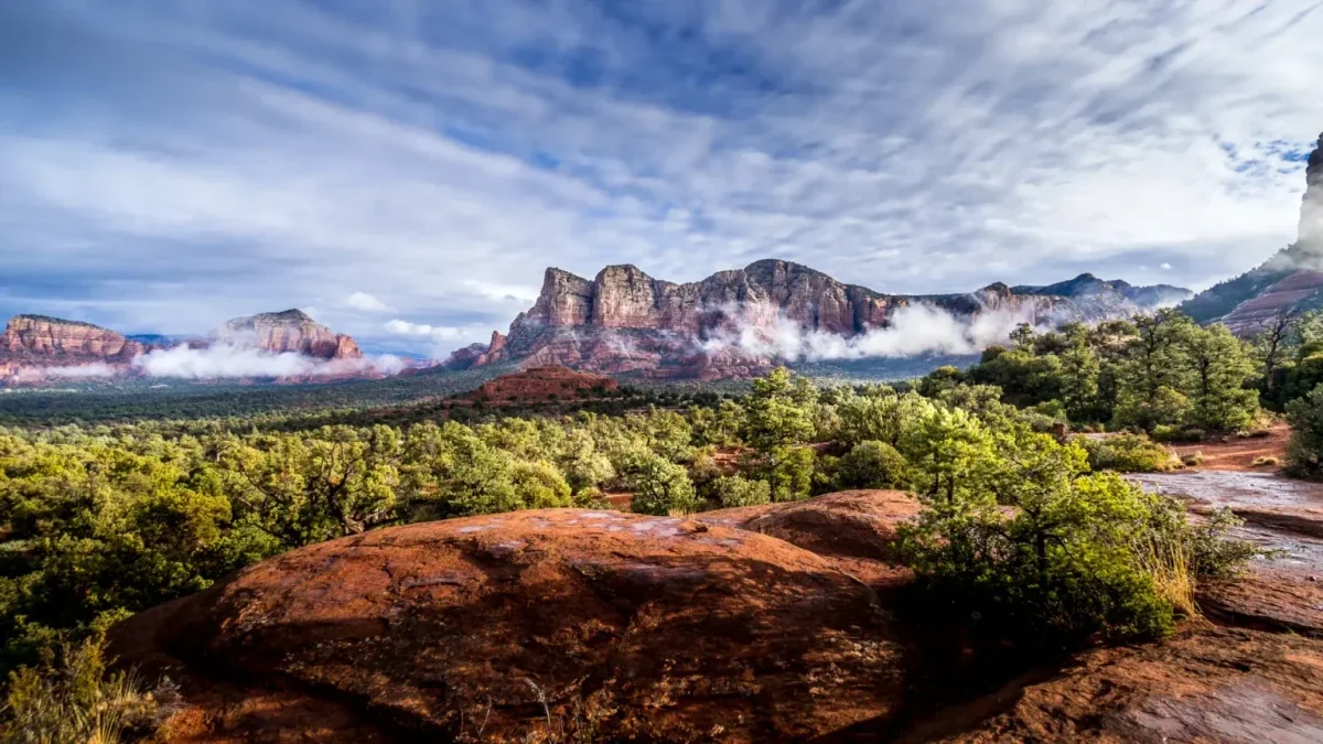 Red rock cliffs with low clouds above green brush and red slickrock