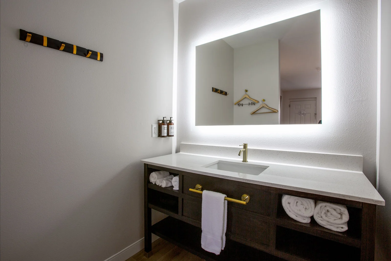 Bathroom vanity at an angle, with the backlit mirror reflecting hangers on the wall hook rack and a towel bar below.