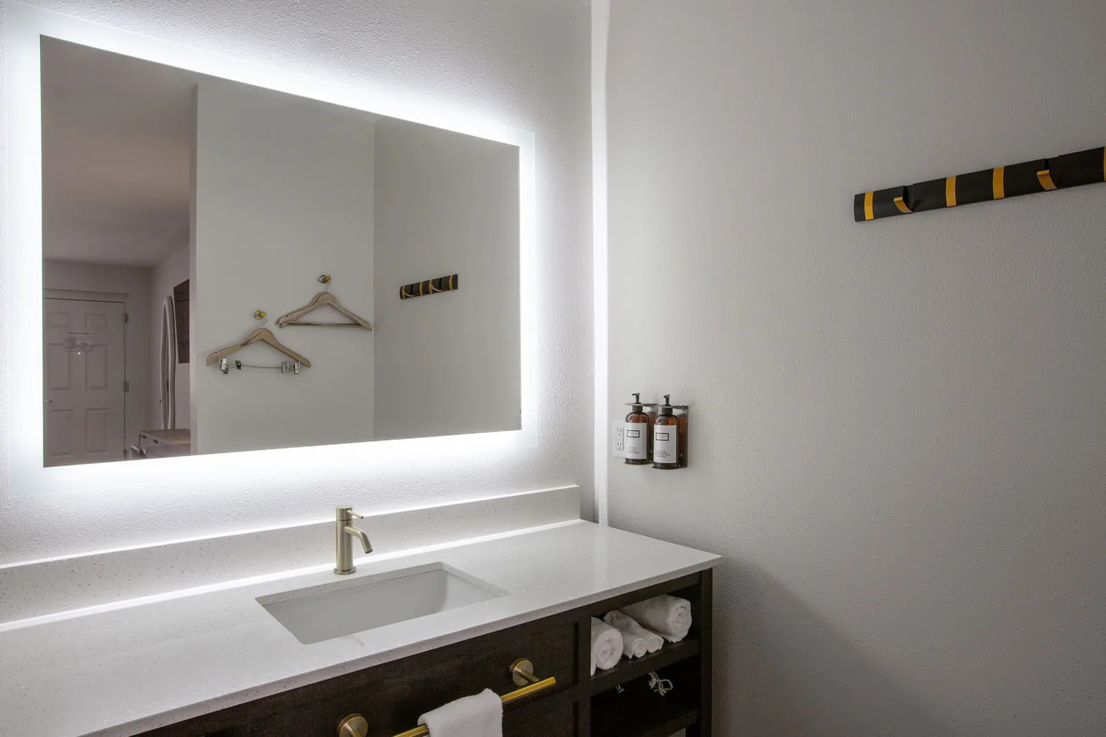 Backlit bathroom mirror with robe hooks and amenity jars on the wall beside it.