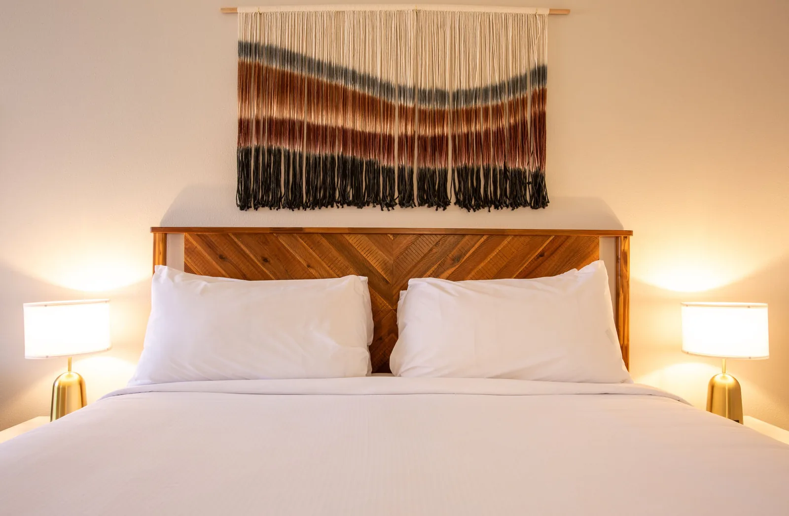 King bed headboard with a woven ombre wall hanging and lit nightstand lamps.