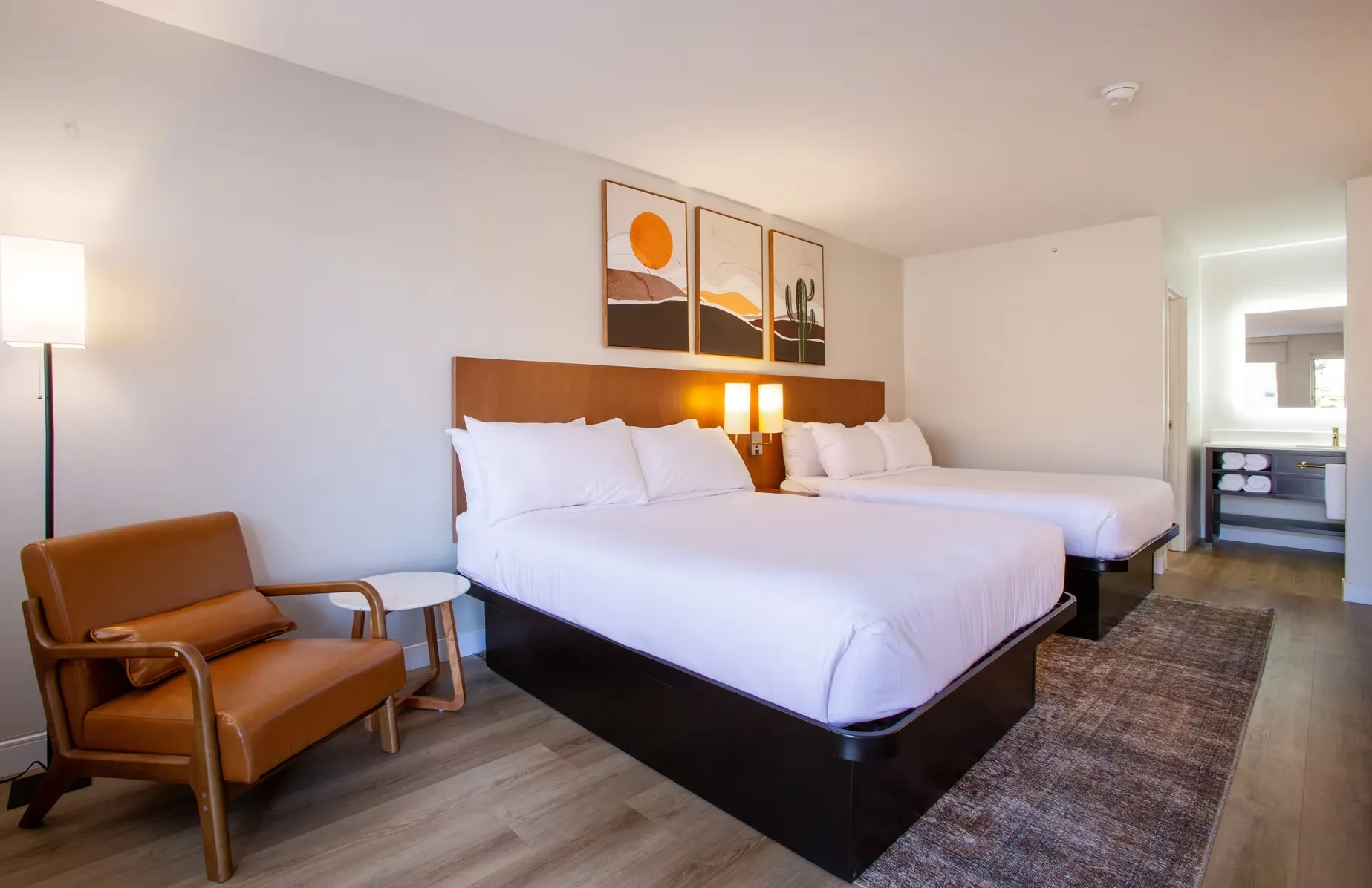 Two queen beds with an orange leather armchair and side table, and a doorway open to an adjoining room.