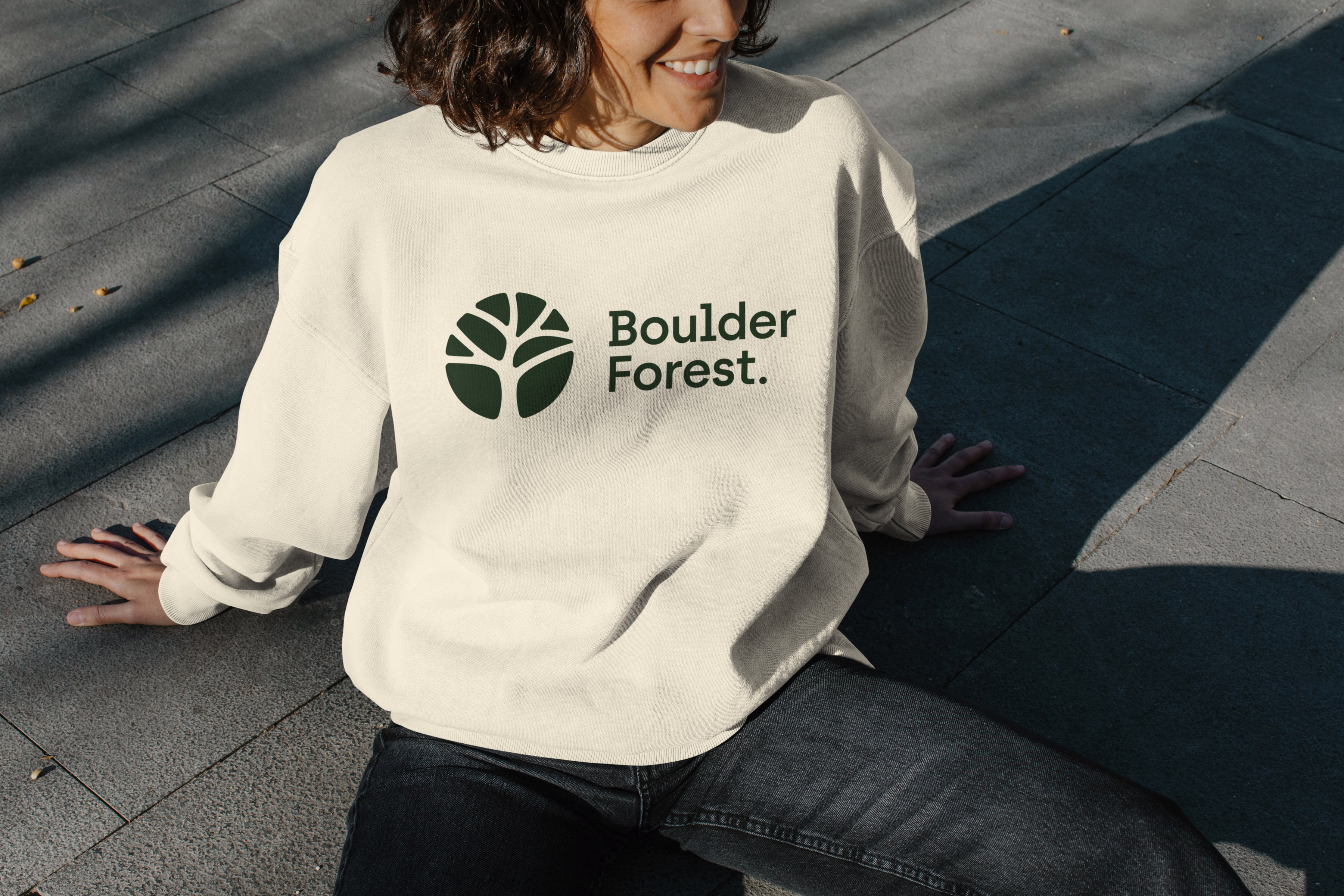Woman in a cream sweatshirt with the dark green BoulderForest logo, sitting on stone steps
