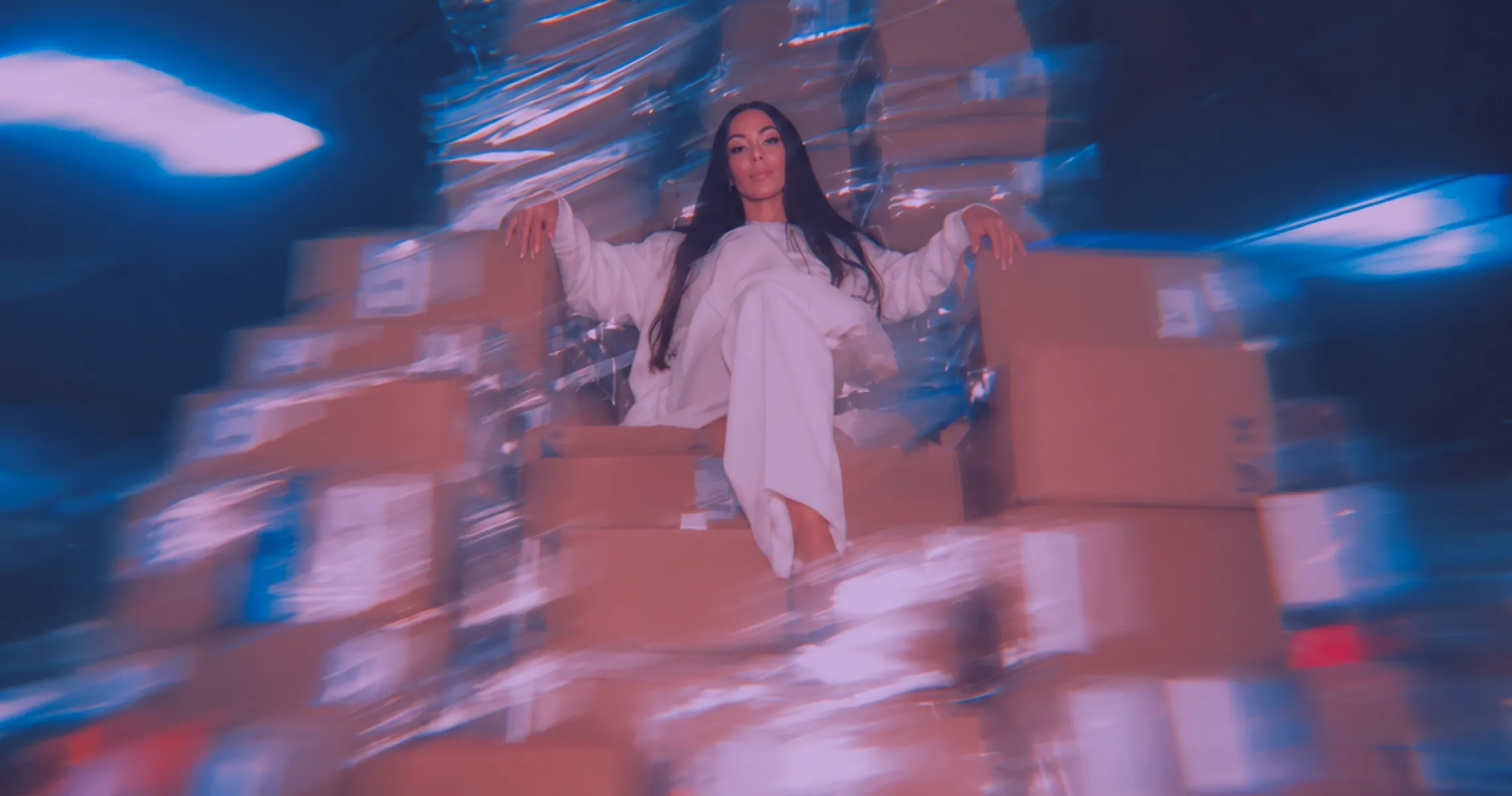 Woman in white loose outfit sitting confidently on a stack of cardboard boxes in a dimly lit, industrial-like setting with reflective plastic wrapping creating a blurred, blue and purple effect.