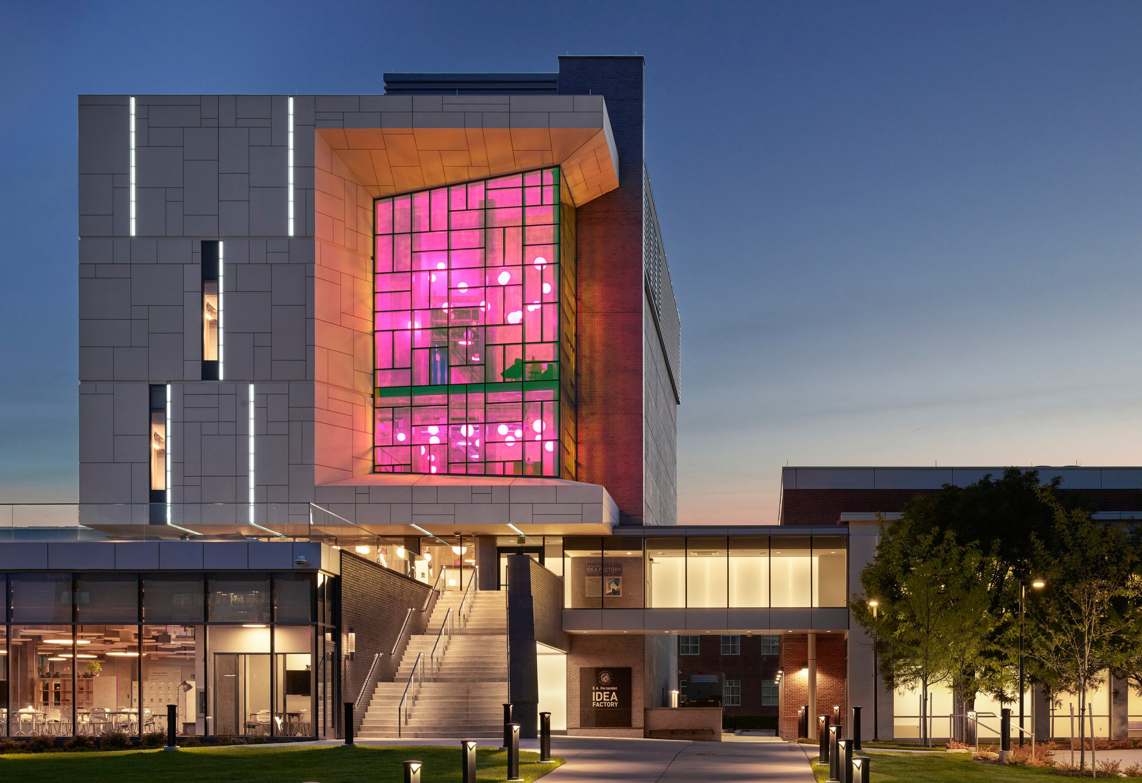 The E. A. Fernandez Idea Factory at dusk, its colored-glass window wall lit magenta above the entrance stair and signage
