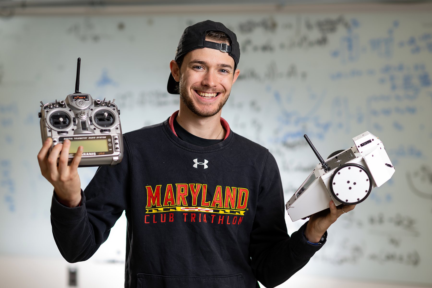 A student in a Maryland Club Triathlon sweatshirt holds a radio transmitter and a wheeled robotics device in a lab