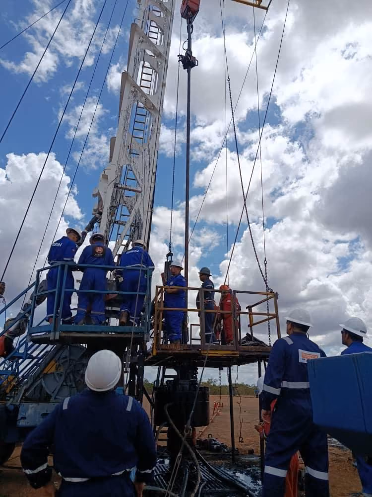 Workers wearing blue uniforms and white helmets operating heavy machinery with cables and a pulley system under a partly cloudy blue sky on a dirt site.