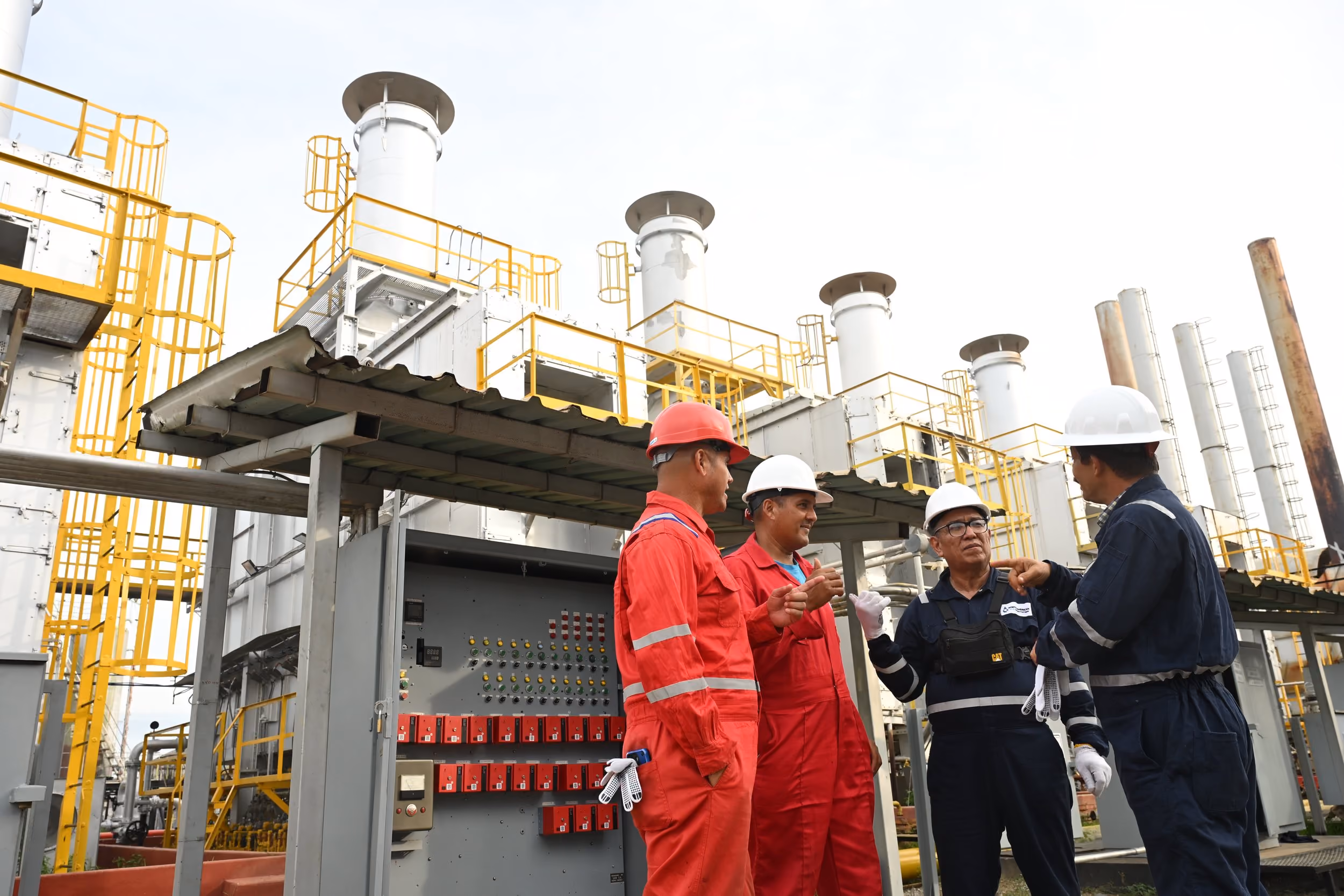 Four industrial workers wearing hard hats and protective coveralls having a discussion by a control panel at a large factory or power plant with metal structures and yellow safety railings.
