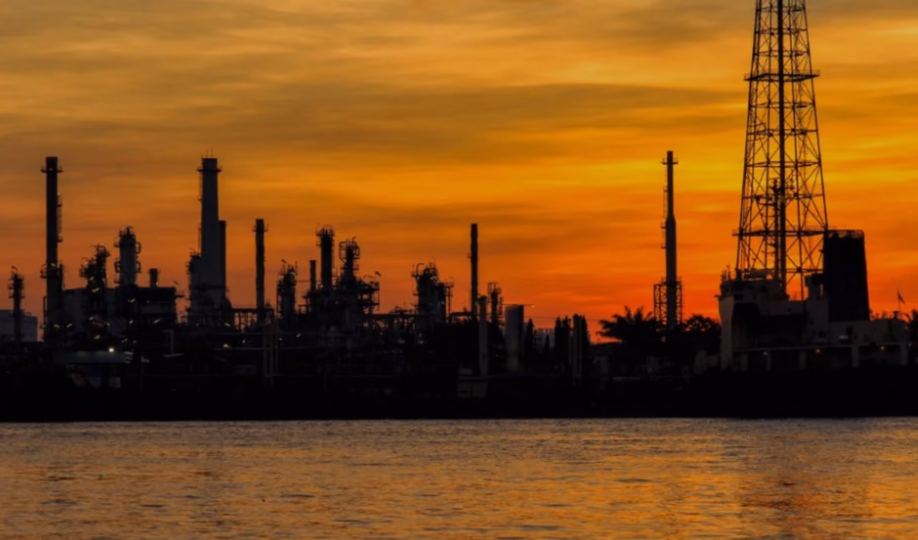 Silhouette of an oil refinery and drilling tower at sunset over the water