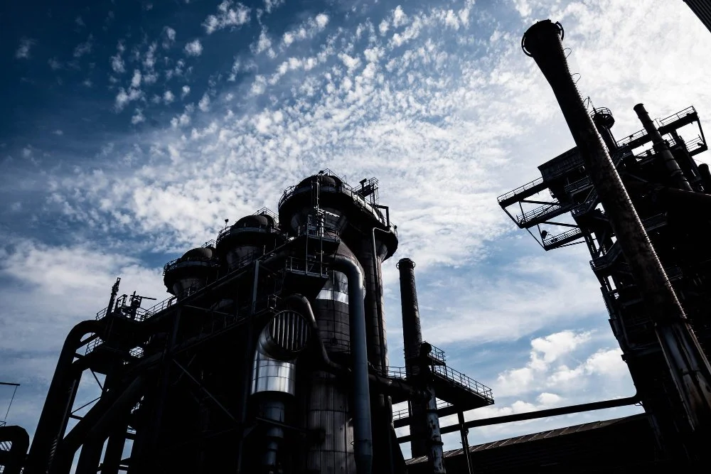 Silhouetted industrial refinery or processing plant with large cylindrical towers, pipes, and platforms set against a partly cloudy blue sky.