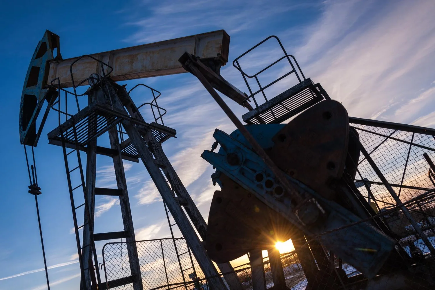 Silhouette of an oil pumpjack against a blue sky with wispy clouds during sunset, with the sun shining through the metal structure near a chain-link fence.