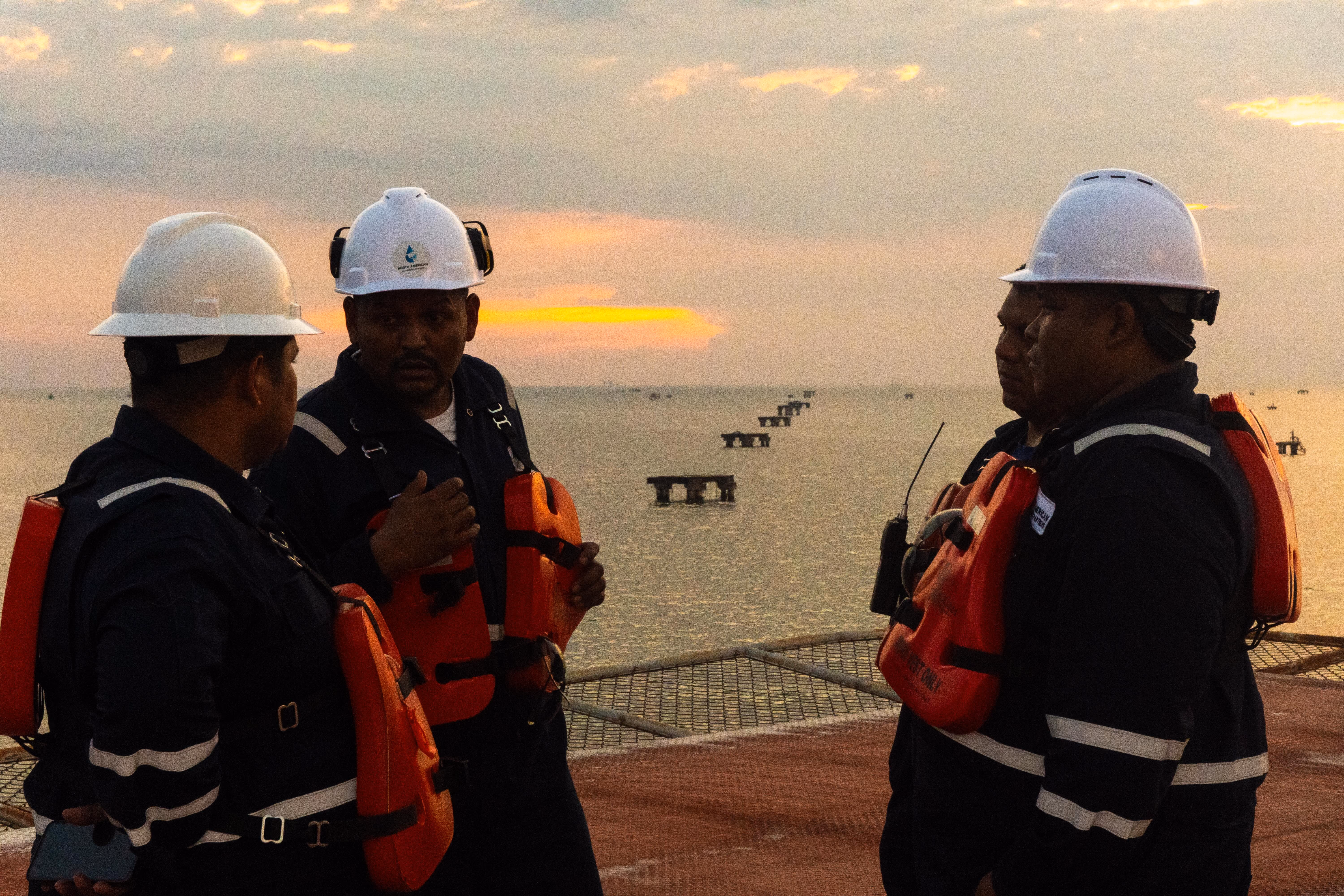 Field workers in hard hats and life vests talking on an offshore platform at sunset