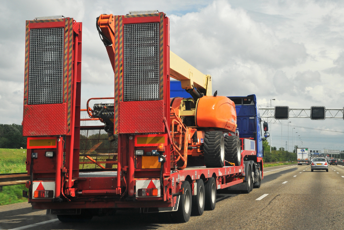Lowboy trailer hauling an orange boom lift and machinery on a highway
