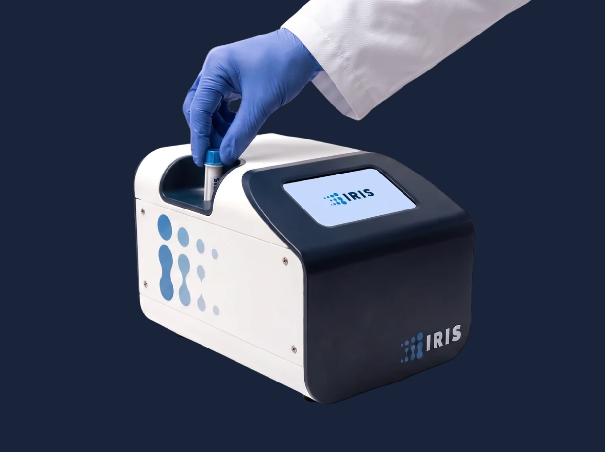 The IRIS TX-100 development prototype: a compact white and navy tabletop analyzer, with a gloved hand inserting a single-use cartridge.