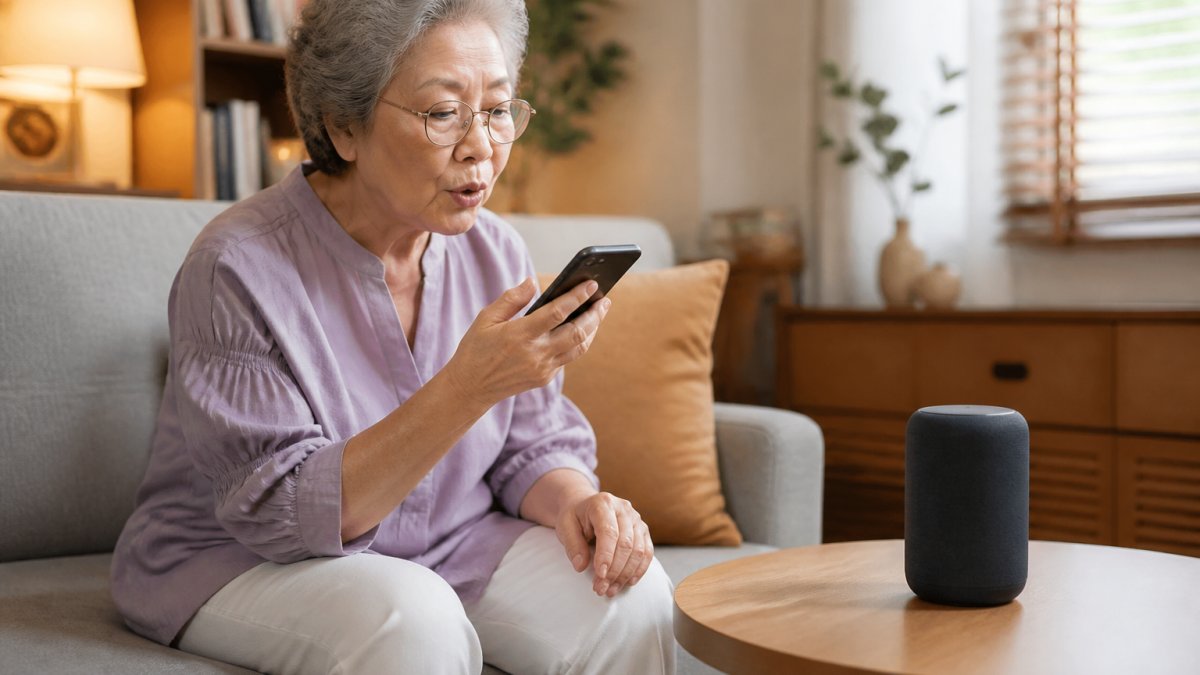 Older adult sitting comfortably at home with a smartphone in hand, speaking to a smart speaker on a nearby table, with warm home lighting in the background