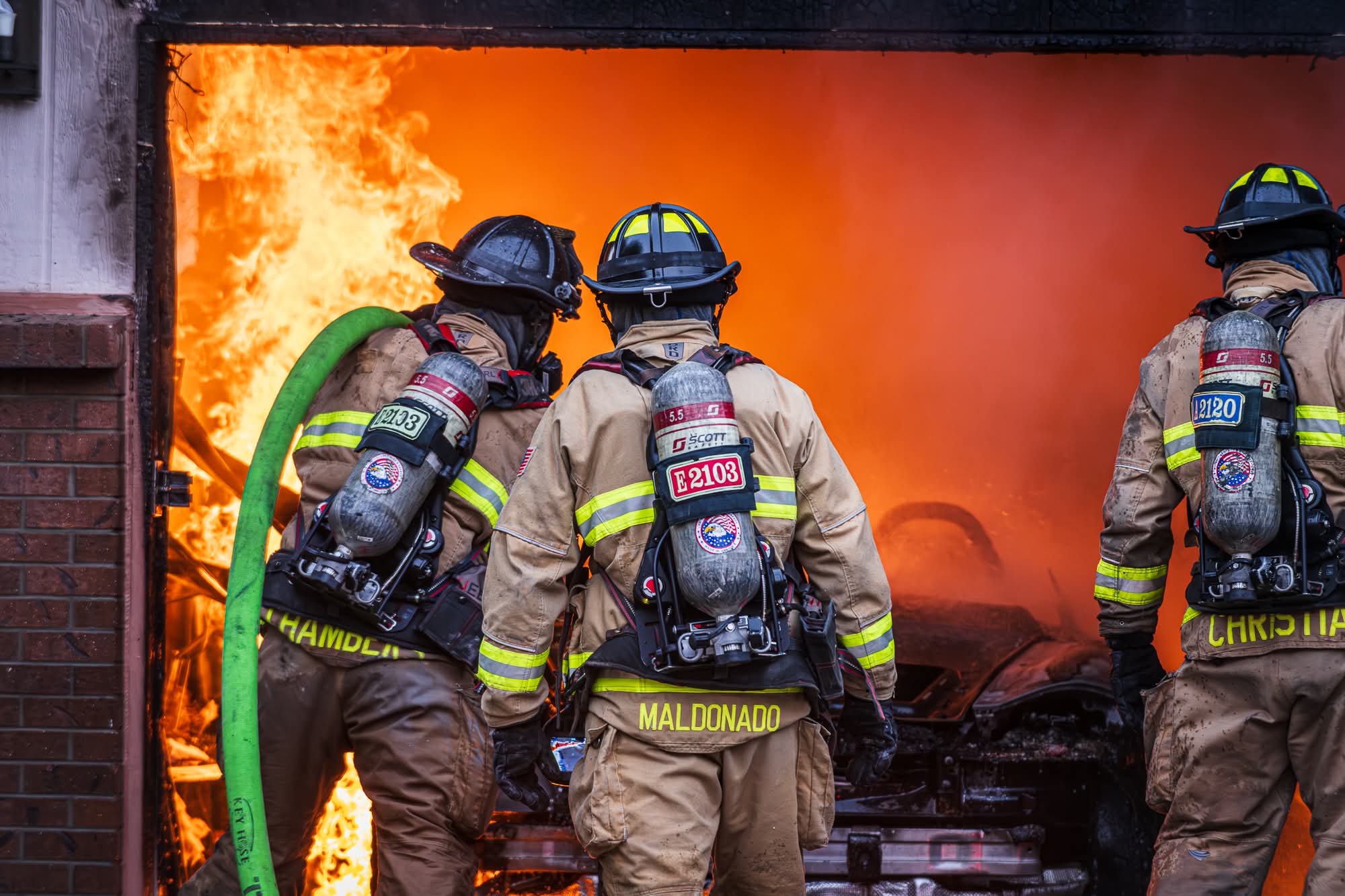 Firefighter kneeling with a hose line in front of a burning house