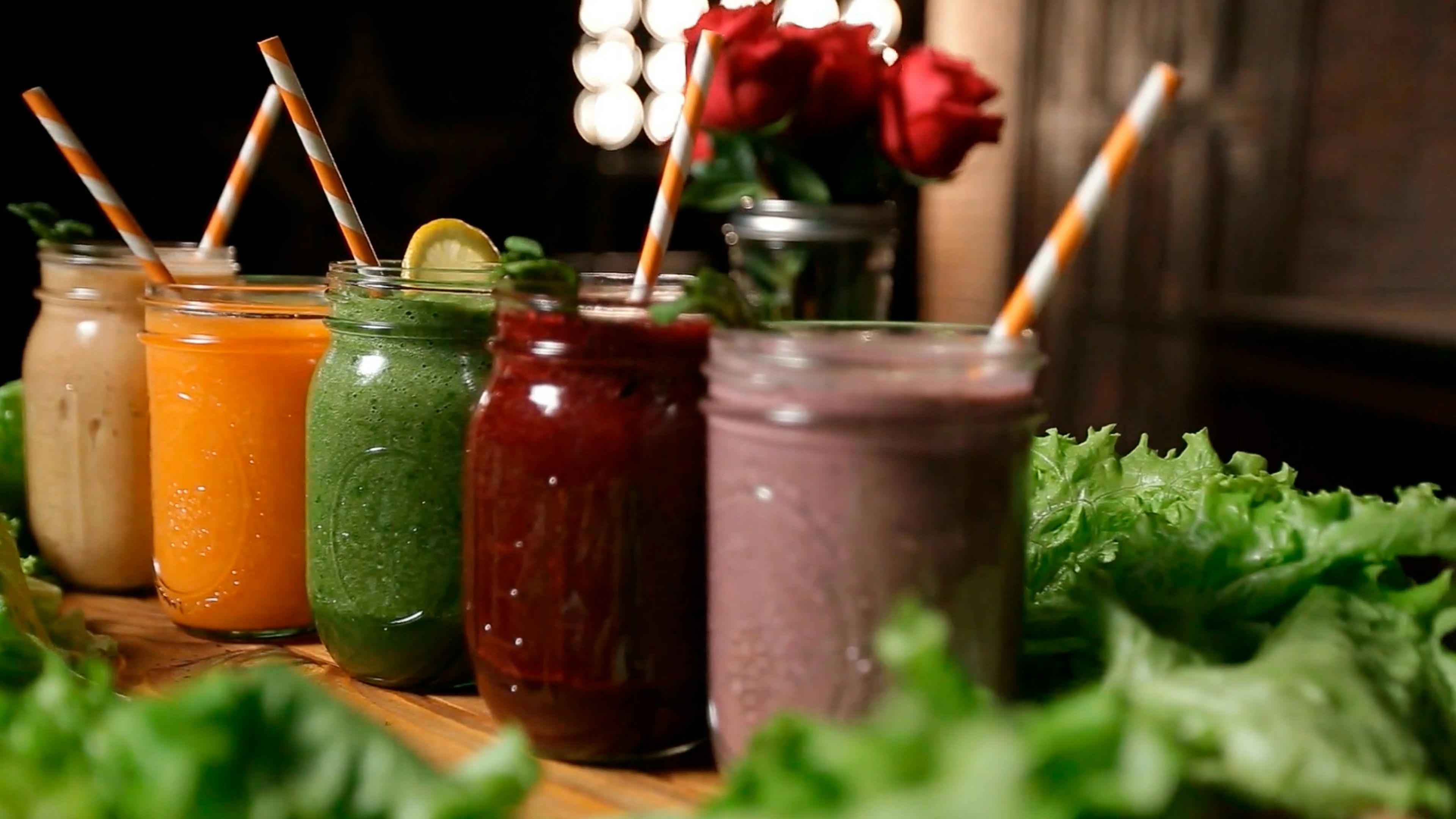 Cinq smoothies de différentes couleurs rangés dans des bocaux en verre avec des pailles rayées orange et blanc, entourés de feuilles de laitue, avec un bouquet de roses rouges flou en arrière-plan.
