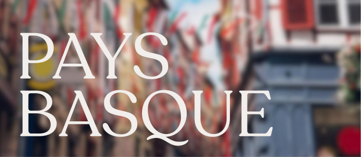 Texte blanc en majuscules indiquant « PAYS BASQUE » sur un fond flou de rue décorée avec de nombreux drapeaux colorés.