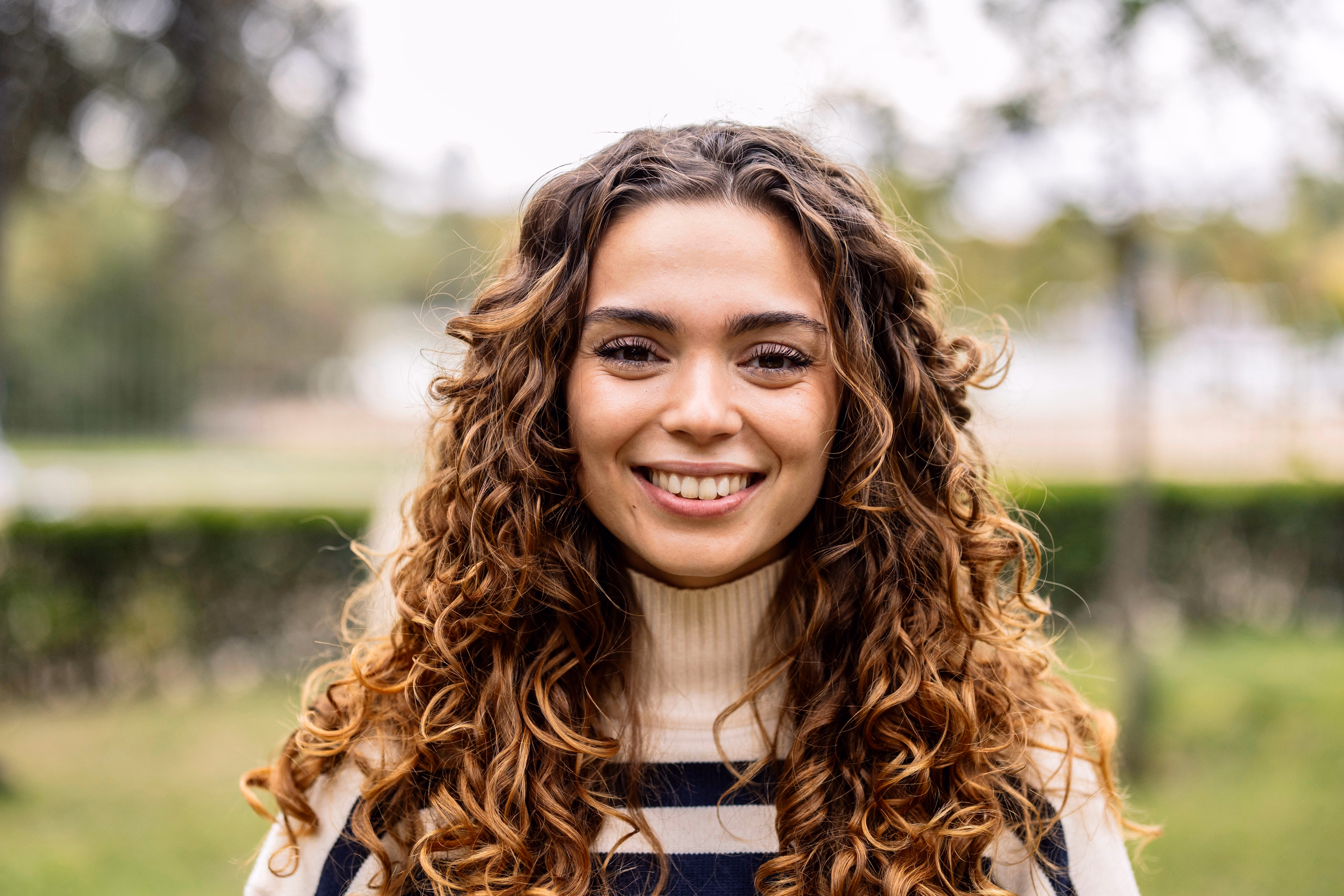 Jeune femme souriante aux longs cheveux bouclés, portant un pull à col roulé beige avec des rayures noires, posant à l'extérieur avec un arrière-plan flou de verdure.