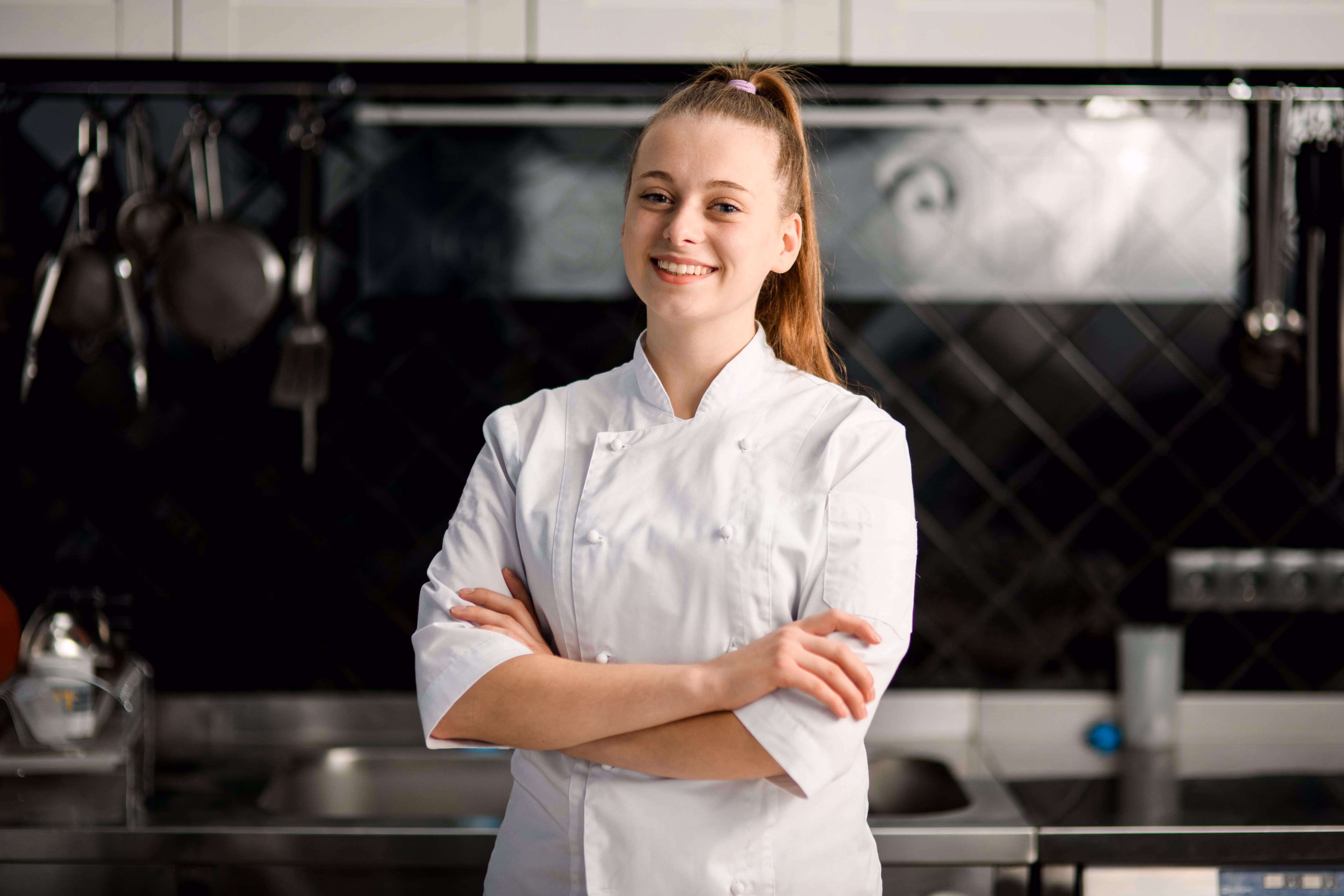 Jeune femme souriante en veste de chef blanc avec les bras croisés dans une cuisine professionnelle avec des ustensiles suspendus en arrière-plan.