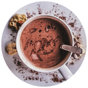 Tasse blanche de chocolat chaud avec une cuillère métallique à l'intérieur, posée sur une assiette avec des éclats de cacao et des morceaux de biscuit autour.