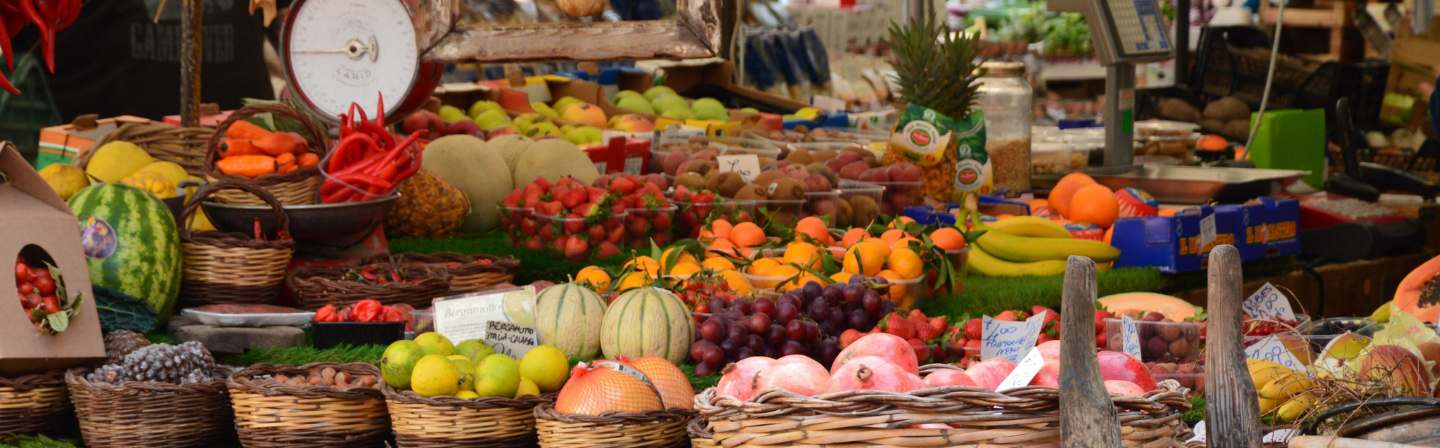 Étalage coloré d'un marché avec divers fruits et légumes, y compris pastèques, fraises, melons, bananes, grenades, carottes, piments rouges, agrumes, raisins, ananas et tomates cerises présentés dans des paniers et sur des tables.
