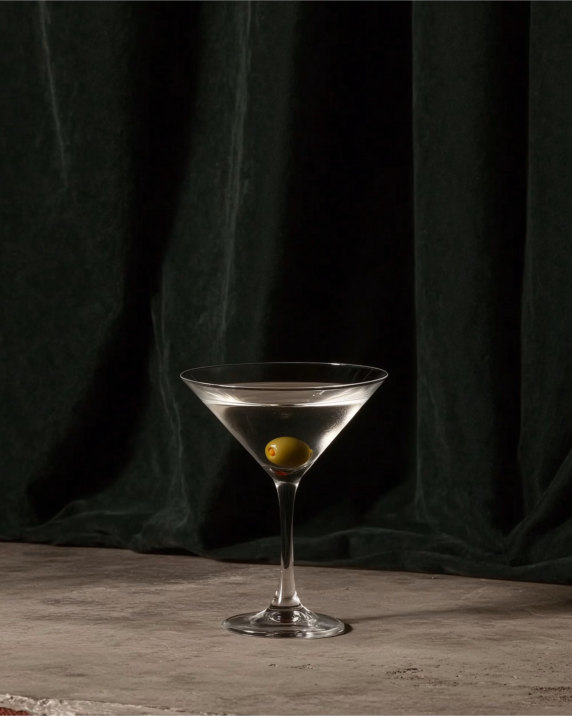 Dirty martini with an olive on stone in front of a dark velvet curtain