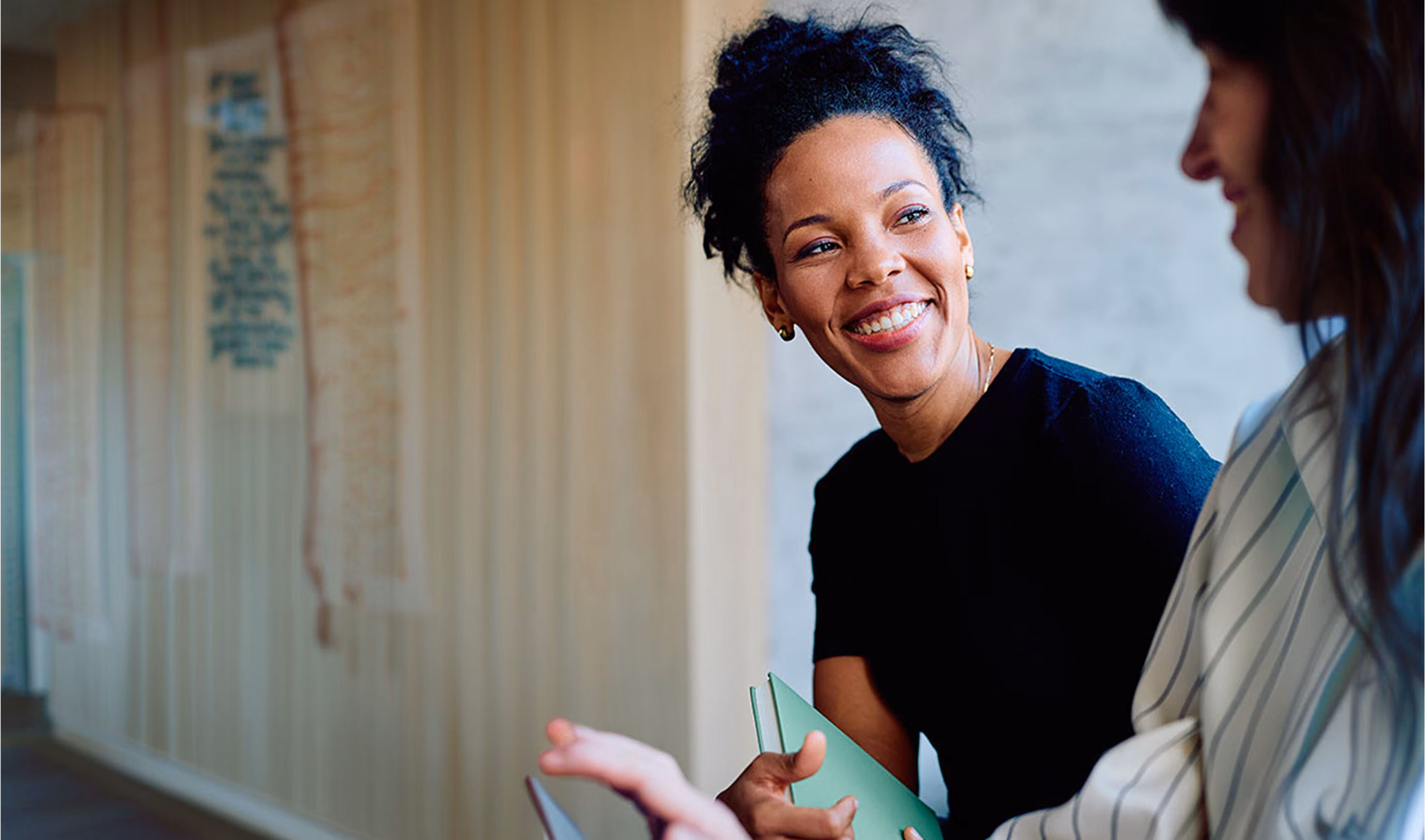 Two women engaged in a friendly conversation, one smiling while holding a green notebook, in a casual office setting with a blurred wall covered in sticky notes in the background.