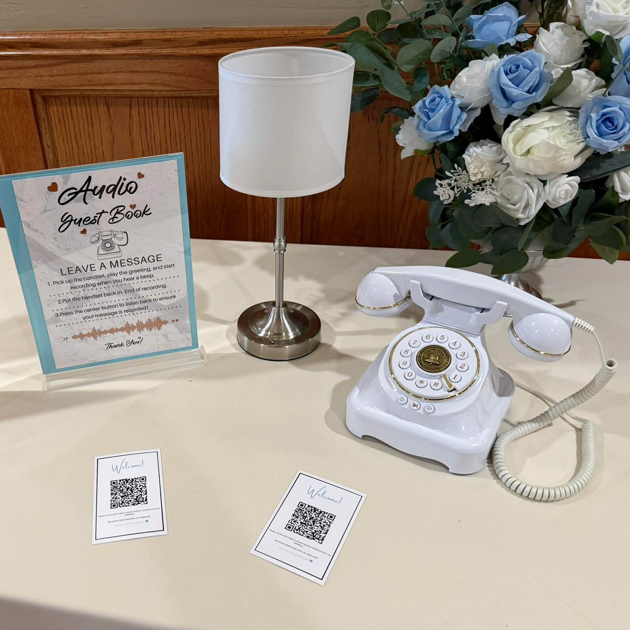 Vintage rotary phone audio guest book display on a welcome table, with instruction card, shuttle bus sign, and floral arrangement.