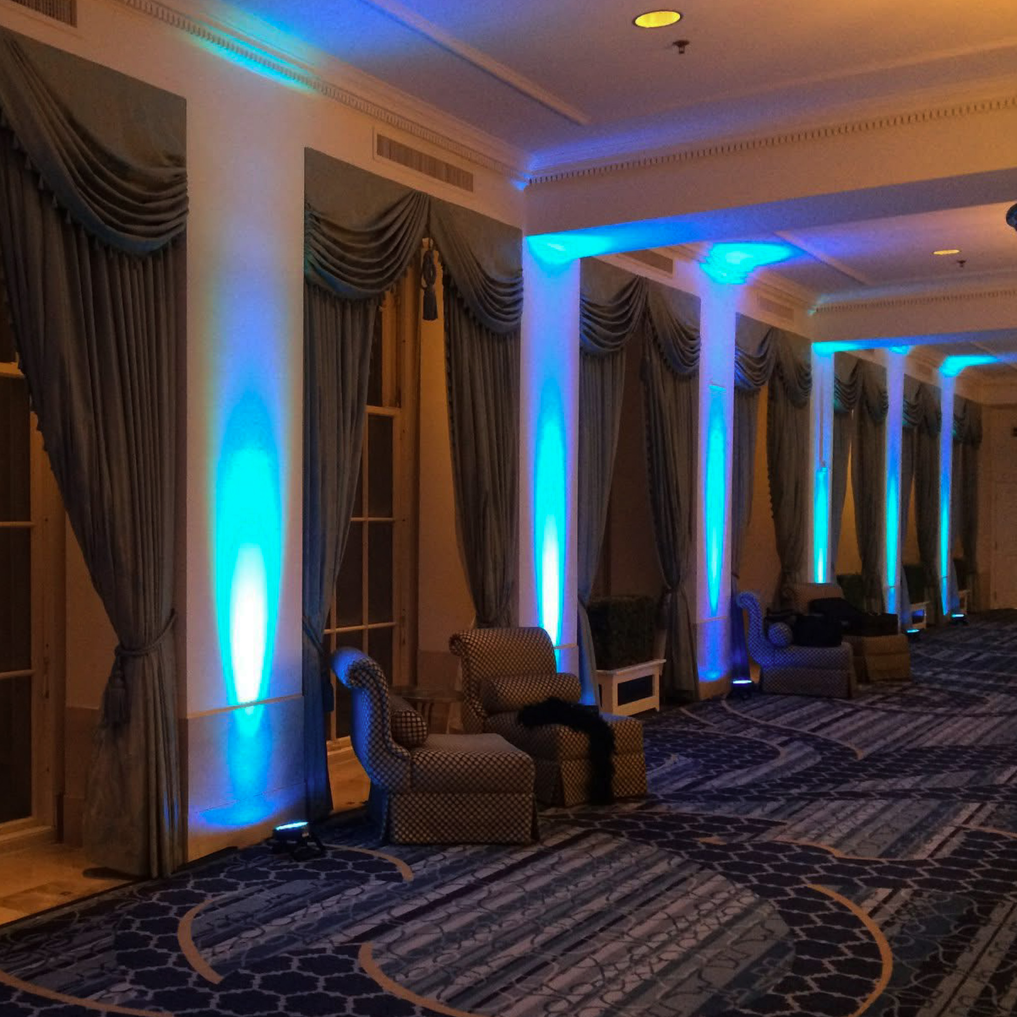 Elegant hotel hallway lined with columns and draped curtains, each lit with blue uplighting along a patterned carpet.