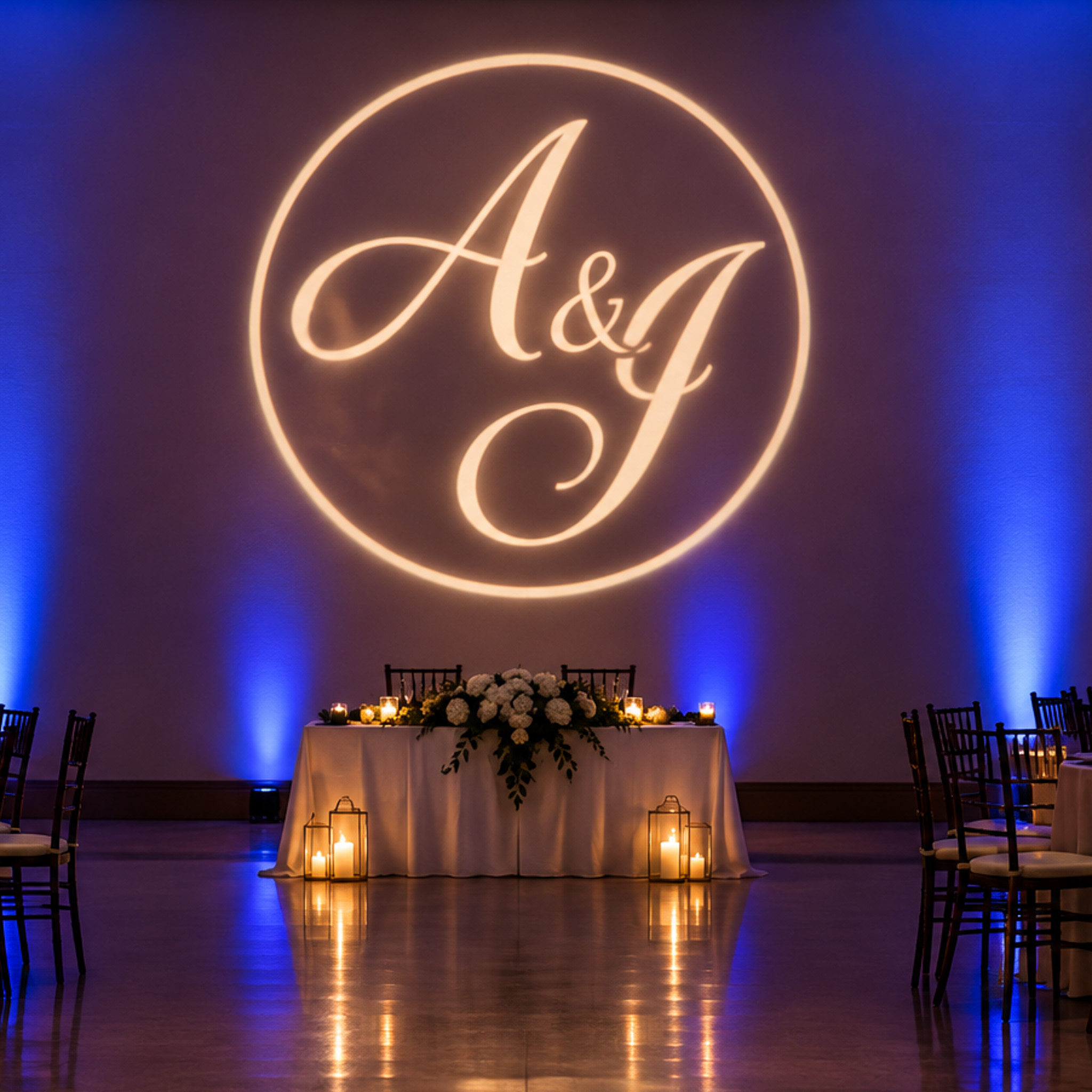 Circular "A&J" monogram light projected on a wall above a candlelit sweetheart table, flanked by blue uplighting and floral arrangements.