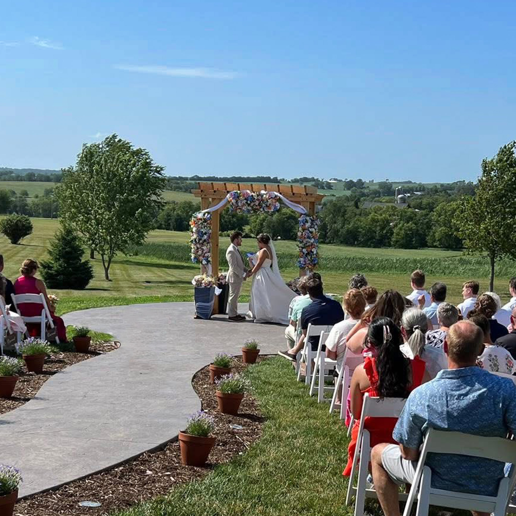 Outdoor wedding ceremony under a floral arch with rolling farmland and trees in the background, guests seated in white folding chairs.
