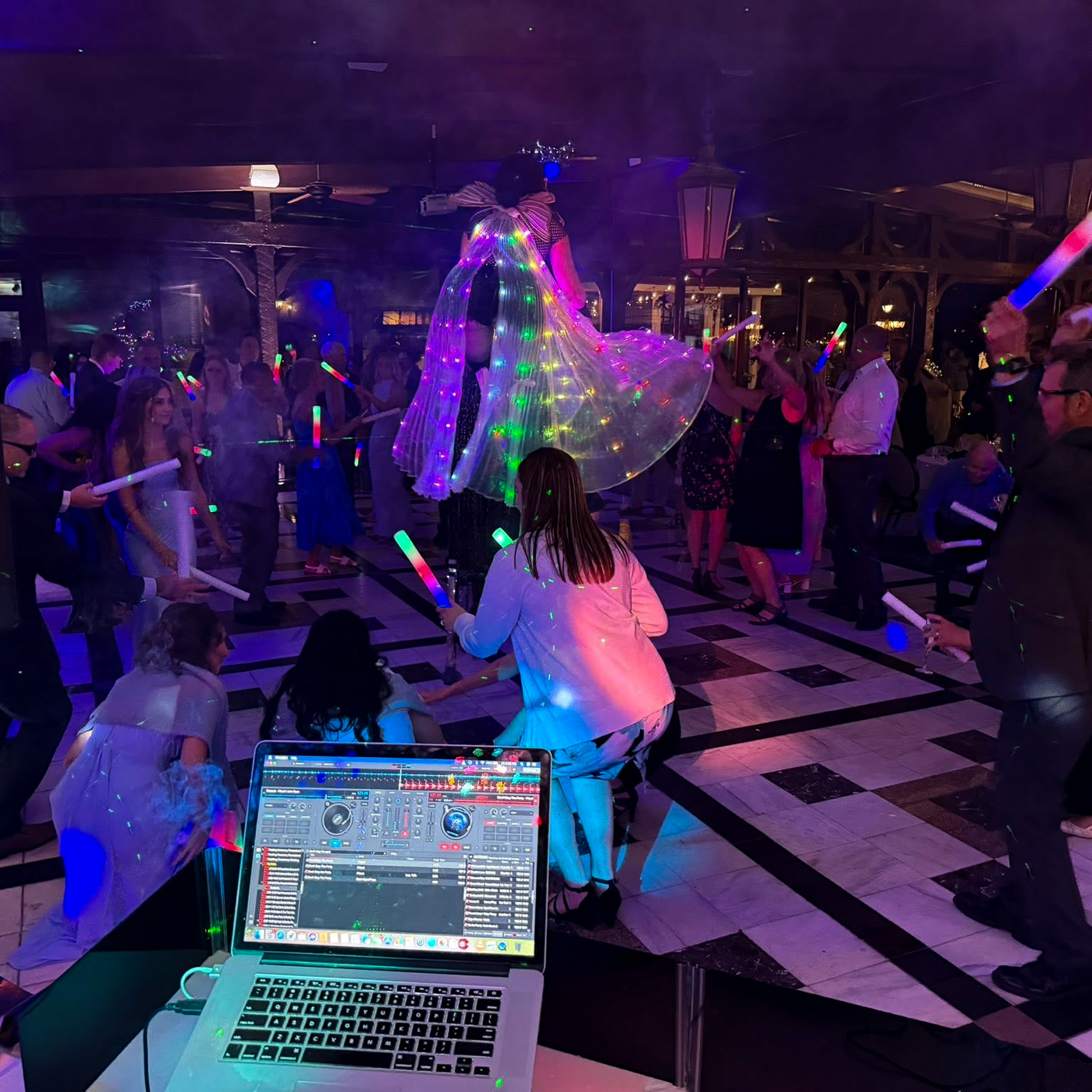 DJ laptop and mixer in the foreground with a lit dance floor behind, guests waving glow sticks around a bride in a light-up cape.